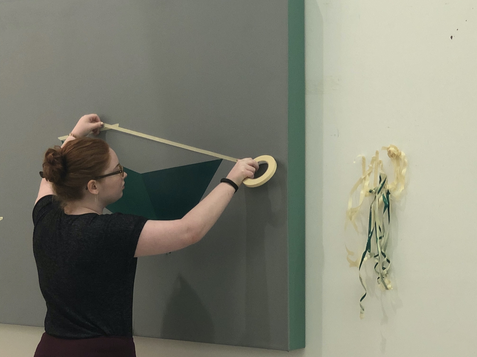 Student stretches masking tape across the surface of a prepared canvas covered in gray and green paint.