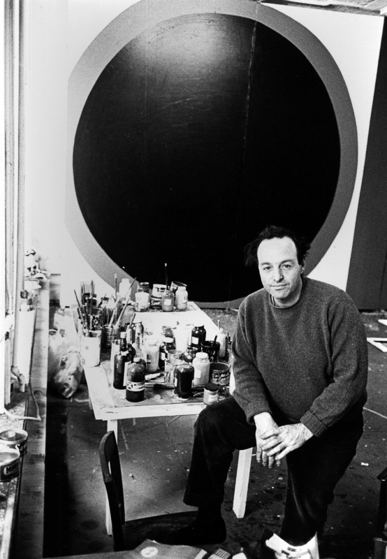 Back and white photo of a white middle aged man with dark hair posing in front of a table covered with paints jars and paint brushes, a large abstract painting hangs in the background
